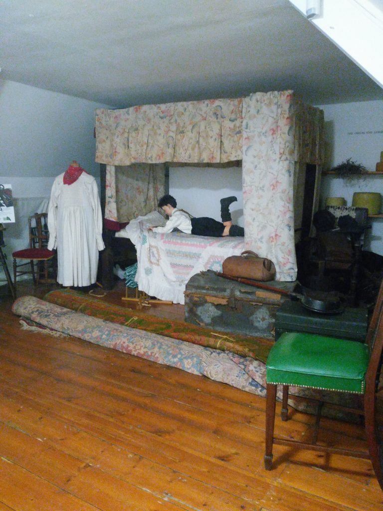 A full view of the room, including the manniquin on the bed, the feet of another visible under the bed, and another showing a dress beside the bed.