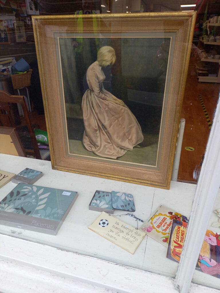 A creepy framed portrait in a charity shop window of a pale-haired woman sitting mournfully in a chair.